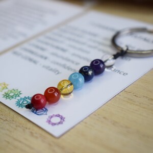 May include: A keychain with seven chakra beads in the colors red, orange, yellow, green, blue, indigo, and violet. The beads are strung on a silver key ring with a black cord.
