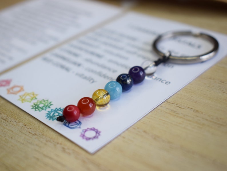 May include: A keychain with seven chakra beads in the colors red, orange, yellow, green, blue, indigo, and violet. The beads are strung on a silver key ring with a black cord.