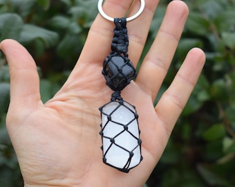 May include: A keychain featuring a black stone and a white crystal, both encased in black macrame. The keychain has a silver ring at the top. The background is a blurred green.