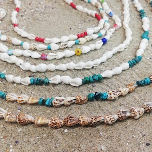 Puede incluir: Una colección de collares de conchas con cuentas de varios colores, incluyendo turquesa, rojo y blanco. Los collares están dispuestos en un patrón circular sobre una superficie arenosa.