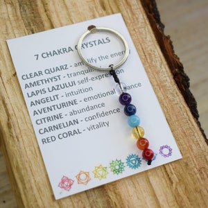 May include: A keychain with seven chakra crystals, each representing a different color of the rainbow. The crystals are strung on a black cord and attached to a silver key ring. The card attached to the keychain lists the crystals and their corresponding chakra meanings.
