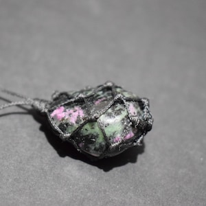 Peut inclure: Un pendentif en pierre précieuse verte et rose avec un cordon noir, enveloppé dans un nœud macramé.