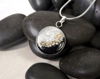 May include: A round pendant necklace featuring a silver-tone snake chain and a circular charm. The charm contains a resin inlay with a swirling white top section, a black bottom section, and a central band of small, textured gold-colored flakes.
