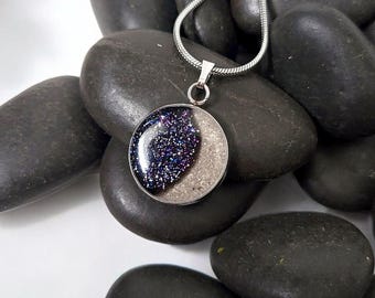 May include: A silver-toned pendant necklace featuring a circular charm with a split design. One half of the charm is filled with a dark blue, glittery material, while the other half is a textured, light beige sand-like surface on a thin chain.