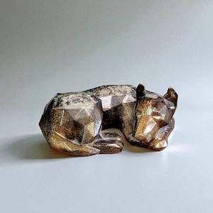 May include: A geometric figurine of a resting dog with a faceted surface. The sculpture features a dark, translucent finish infused with shimmering gold and bronze glitter particles throughout the material, creating a reflective, multi-dimensional appearance.