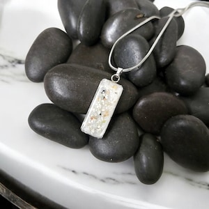 May include: A rectangular pendant necklace with a silver chain. The pendant is clear with small white, beige, and black inclusions. The necklace rests on a bed of smooth, dark gray stones.