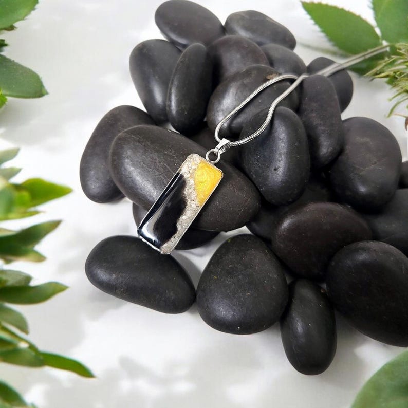 May include: A silver chain necklace with a rectangular pendant featuring a black and white agate stone with yellow accents. The pendant is set against a background of black stones.