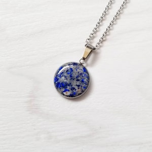 May include: A circular pendant necklace featuring a silver-toned metal setting and chain. The pendant contains a smooth, resin-like inlay filled with small, vibrant blue flecks and light gray, sandy-textured particles throughout the surface.