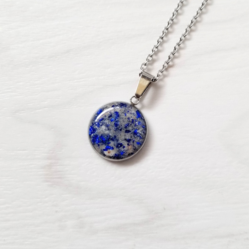 May include: A circular pendant necklace featuring a silver-toned metal setting and chain. The pendant contains a smooth, resin-like inlay filled with small, vibrant blue flecks and light gray, sandy-textured particles throughout the surface.