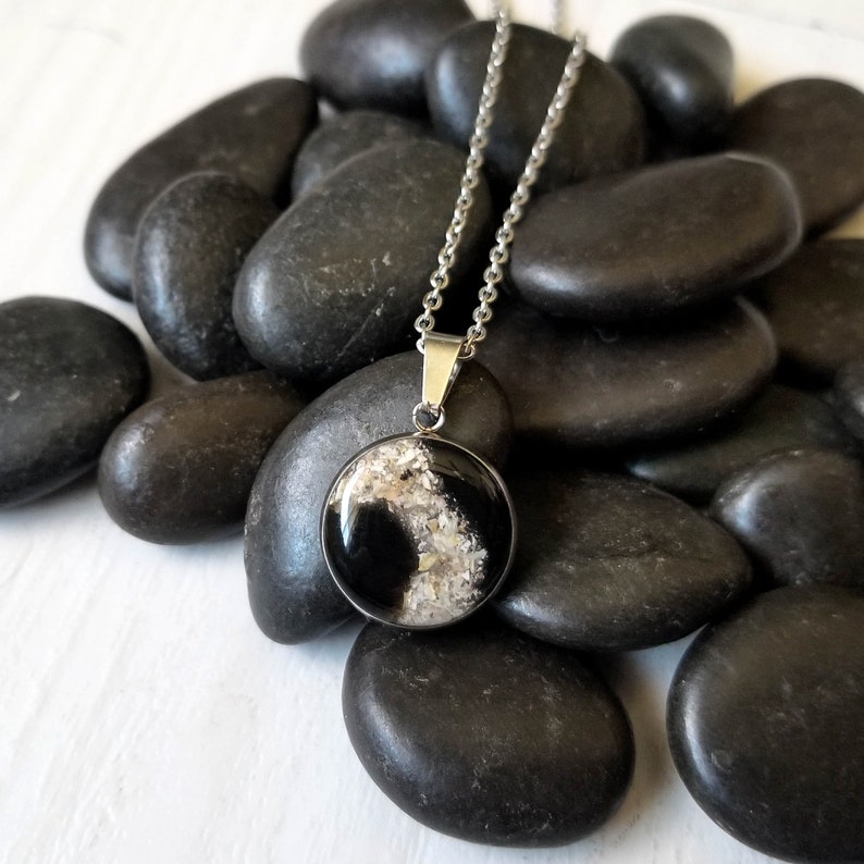 May include: A circular pendant necklace featuring a silver-toned metal setting. The pendant contains a glossy black resin inlay accented with a textured, light-colored granular material that creates a distinct contrast against the dark background.