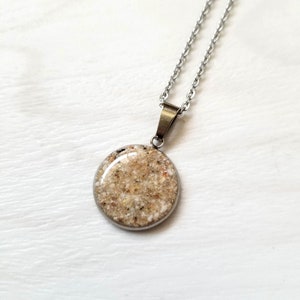 May include: A silver-toned metal pendant necklace featuring a circular charm filled with textured, multi-colored sand grains under a smooth, clear, domed resin finish, suspended from a matching silver-toned link chain.
