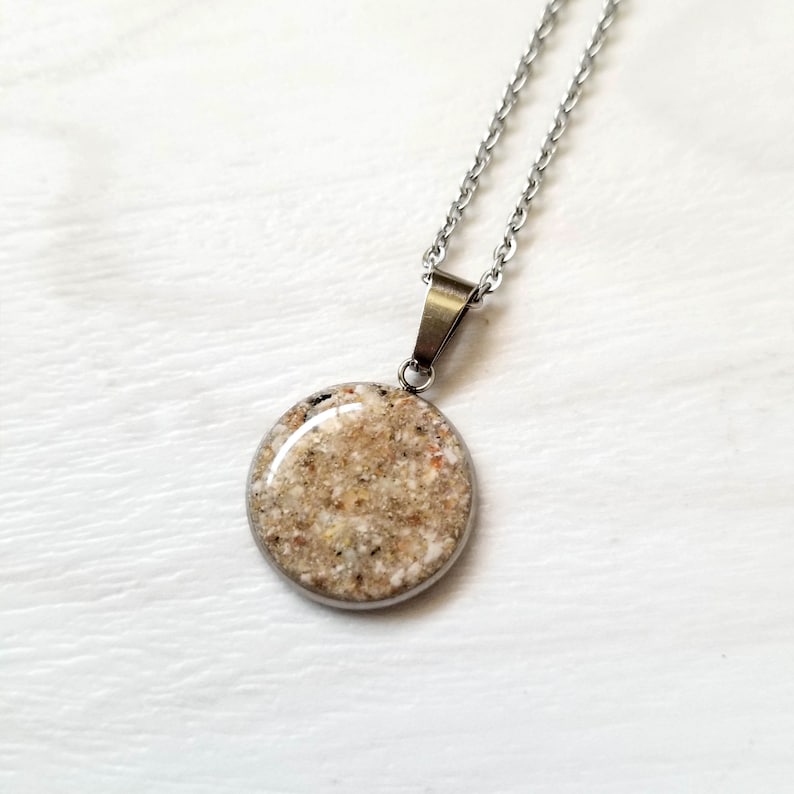 May include: A silver-toned metal pendant necklace featuring a circular charm filled with textured, multi-colored sand grains under a smooth, clear, domed resin finish, suspended from a matching silver-toned link chain.