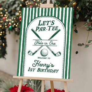 May include: A rectangular sign with a white background and dark green vertical stripes on the sides. The center features a dark green arched frame containing the text LET'S PAR-TEE, Hole in One, WELCOME TO, Henry's 1ST BIRTHDAY, and golf clubs.