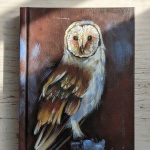 Può includere: Dipinto dettagliato di un barbagianni su una piccola tela. Il gufo ha una faccia bianca a forma di cuore, piume marroni e dorate ed è appollaiato su una superficie blu su uno sfondo marrone scuro. L'opera è firmata in basso a destra.