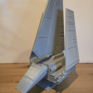 Puede incluir: Modelo gris impreso en 3D de una nave espacial, con alas y cuerpo detallados. El modelo tiene una superficie texturizada y está sobre un fondo neutro. El diseño incluye paneles intrincados y un emblema circular.