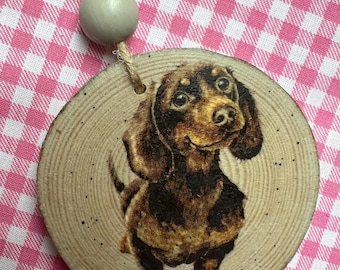 May include: A wooden round ornament with a painted image of a brown and black dachshund dog. The ornament has a twine string with a light gray bead.