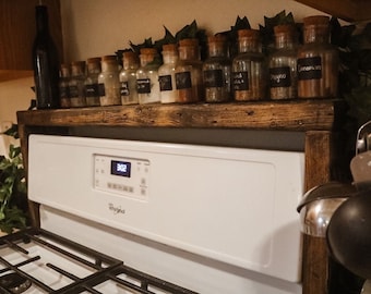 May include: A rustic wooden spice rack with glass jars filled with various spices, each labeled with a black chalkboard label. The rack sits above a white Whirlpool oven. Decorative greenery adds a natural touch.