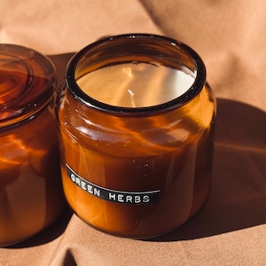 May include: Two amber glass candle jars with lids. The candle in the foreground has a white label with the text "GREEN HERBS".