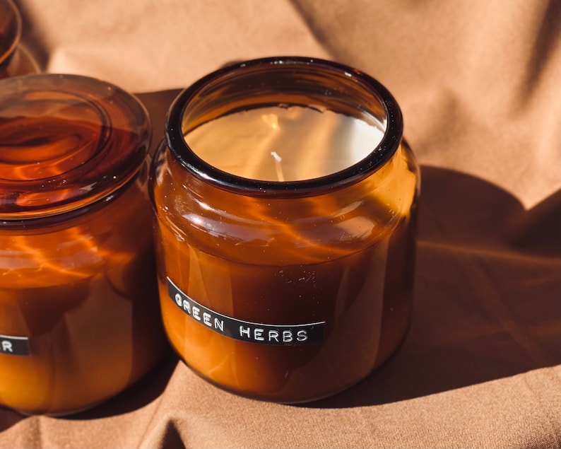 May include: Two amber glass candle jars with lids. The candle in the foreground has a white label with the text "GREEN HERBS".