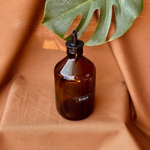 Op de afbeelding: Een bruine glazen fles met een zwarte giettuit en een etiket met de tekst "Suiker".