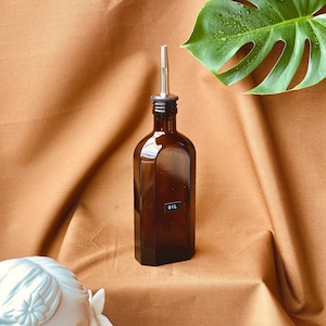 May include: A brown glass bottle with a silver spout and a black label that says "OIL". The bottle is sitting on a brown fabric surface.