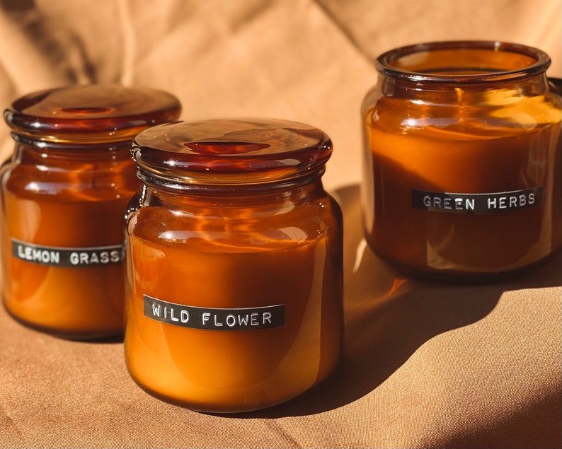 May include: Three amber glass candle jars with black labels. The labels read "Lemon Grass", "Wild Flower", and "Green Herbs".