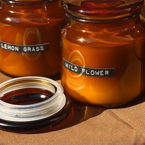 May include: Three amber glass candle jars with black labels. The labels read "Lemon Grass", "Wild Flower", and an unreadable label. The jars are sitting on a brown surface.
