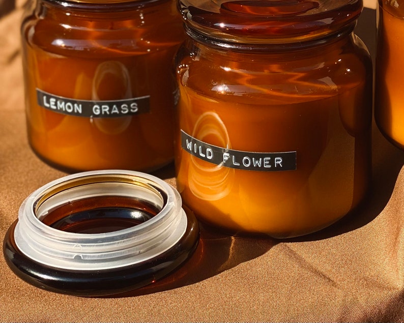 May include: Three amber glass candle jars with black labels. The labels read "Lemon Grass", "Wild Flower", and an unreadable label. The jars are sitting on a brown surface.