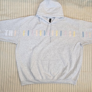 May include: A light gray hoodie with a front pocket and a hood. The sleeves feature the text "I ❤️ YOU THIS MUCH" in white, with colorful vertical stripes. The hoodie is laid flat on a patterned surface.