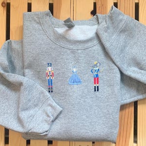 May include: A gray sweatshirt with an embroidered design of a nutcracker, a ballerina, and a mouse. The design is in shades of blue, red, and white.