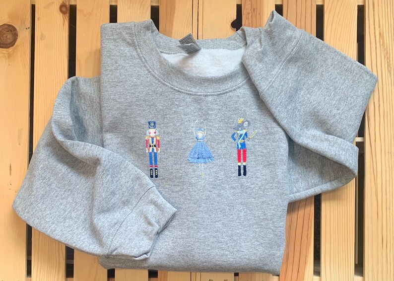 May include: A gray sweatshirt with an embroidered design of a nutcracker, a ballerina, and a mouse. The design is in shades of blue, red, and white.