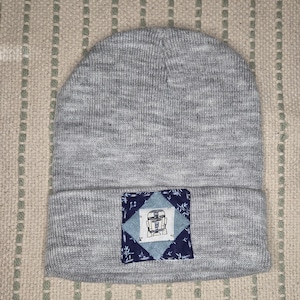 May include: A gray beanie with a square patch featuring a robot design. The patch has a blue and white geometric pattern with a light blue center. The beanie is made of a soft, knitted material.