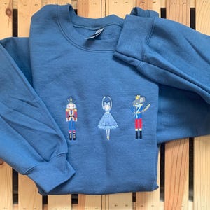 May include: A blue sweatshirt with an embroidered design of the Nutcracker, a ballerina, and a prince. The design is in shades of red, white, blue, and green.