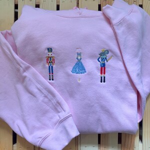 May include: Pink hoodie with embroidered nutcracker, ballerina, and mouse characters.
