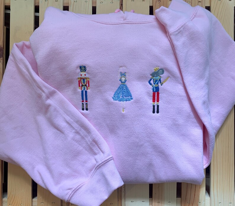 May include: Pink hoodie with embroidered nutcracker, ballerina, and mouse characters.