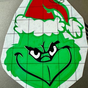 May include: A green Grinch face with a black outline and white eyes wearing a red Santa hat with a white trim. The image is on a white background with a grid pattern.