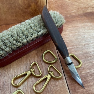 May include: A set of five gold-colored metal key rings with a small wooden handle knife with a metal blade. The knife is lying on a wooden surface next to a brown brush with white bristles.