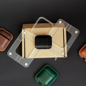 May include: An overhead shot of a black surface with a brown cardboard box, clear plastic templates, and leather AirPod cases in brown and green. A black AirPod case sits on the box.