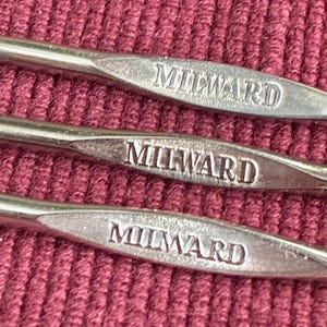 Pode incluir: Três agulhas de crochê de metal na cor prata dispostas em paralelo. Cada agulha apresenta uma seção de cabo achatada com o nome da marca MILWARD gravado em letras maiúsculas. As agulhas possuem um acabamento liso e reflexivo.