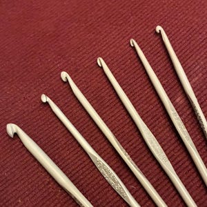 Puede incluir: Primer plano de siete ganchillos plateados de diferentes tamaños, dispuestos en diagonal sobre un fondo burdeos. Los ganchillos tienen extremos curvados y mangos largos. Se puede leer "Made in England" en los ganchillos.
