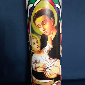 May include: White candle with a colorful stained glass design featuring a saint holding a baby. The design includes the text "SAN ANTONIO".
