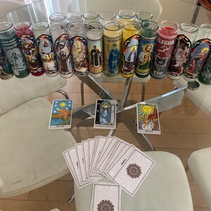May include: A glass table with a selection of candles and tarot cards. The candles are in glass jars with colourful labels featuring religious figures and saints. The tarot cards are spread out on the table, including the Star, The High Priestess, and The Sun cards.