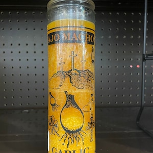 May include: A yellow candle with a black and white label that says "AJO MACHO GARLIC". The label also features a garlic bulb, three crosses, and a clover.