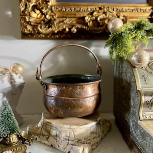 May include: A small, antique copper cauldron with a handle. The pot is round with a dark interior and sits on a rustic, wooden base. The copper has a warm, aged patina. Decorative elements are in the background.