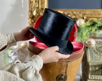 May include: A black top hat with a smooth, dark finish and a wide, structured brim. The hat features a matching black ribbon band around the base of the crown. It is held above a tan, oval-shaped storage case with a red interior lining.