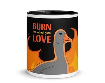 May include: A white ceramic mug with a black rim. The mug features a black background with a gray goose in flames. The text "BURN for what you LOVE" is printed in orange on the mug.