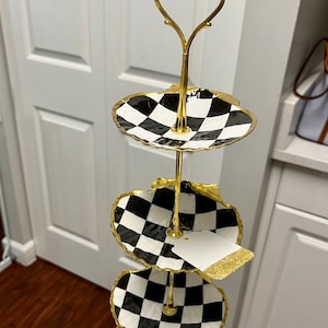 Pode incluir: Um suporte de servir de três níveis com alça e suporte dourados. Os três níveis ovais apresentam um padrão xadrez em preto e branco com acabamento dourado. Uma etiqueta em branco com glitter dourado está anexada.