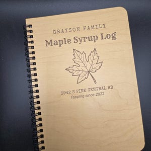 May include: A wooden spiral bound journal with the text "Grayson Family Maple Syrup Log" and a maple leaf design. The address "3942 S Pine Central Rd" and the text "Tapping since 2022" are also engraved on the cover.