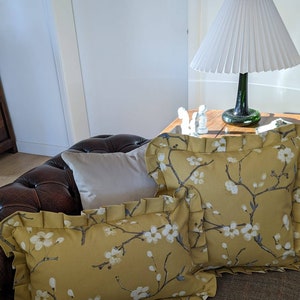 May include: Two mustard yellow throw pillows with white floral patterns and ruffled edges on a brown leather sofa.