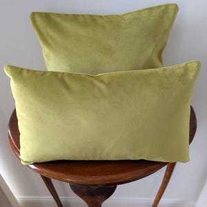May include: Two lime green rectangular cushions. The cushions are made of a soft, velvety material and are placed on a dark wooden side table. The cushions have a simple, elegant design, perfect for adding a pop of colour to any room.